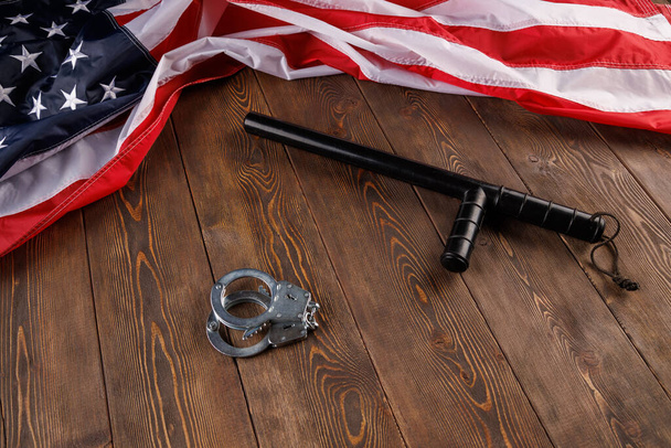 серебряные металлические наручники и полицейская дубинка возле флага США на деревянной поверхности - Фото, изображение