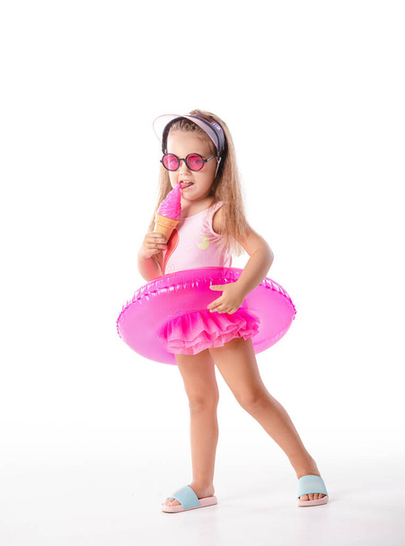 Happy positive little girl stands with inflatable swim ring wears pink swimwear and sunglasses ready for a swim isolated on white background. - Foto, immagini