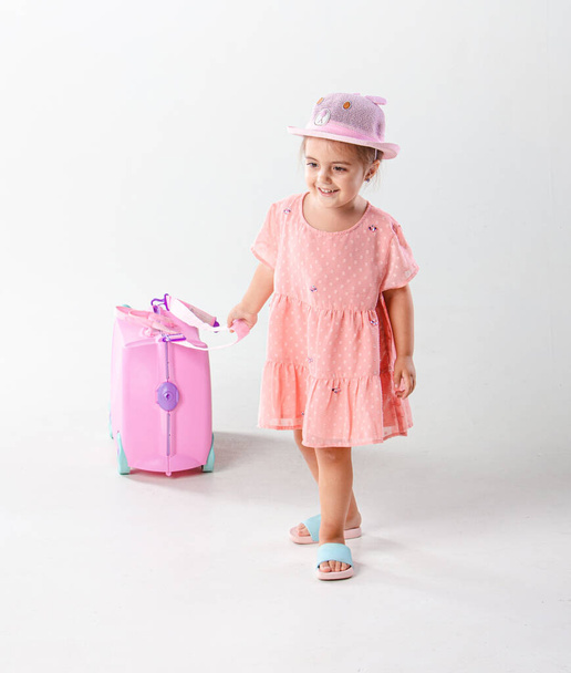 Pretty little kid girl 4-5 years old in pink dress and hat Hold  pink suitcase isolated on white background. Passenger traveling abroad on weekends getaway. Air flight journey concept.  - Фото, зображення