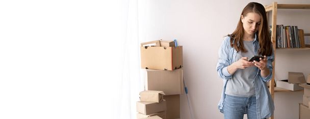serious young woman is moving, standing among cardboard boxes, using a smartphone, moving day and delivery call, banner, space for text - Fotó, kép