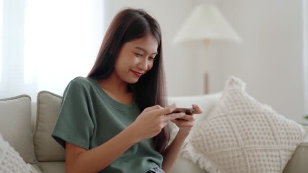Happy young asian woman relax on comfortable couch at home, playing online gaming on smartphone, smiling girl use cellphone chatting, browse wireless internet on gadget, shopping online from home - Séquence, vidéo