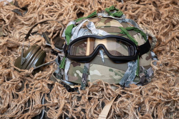 Military camouflage safety helmet and anti-fog protective tactical goggles and noise canceling headphones on camouflage background, military armor, military uniform - Photo, Image