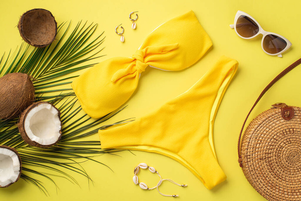 Concepto de vacaciones de verano. Foto vista superior de traje de baño amarillo ronda bolso de ratán elegantes gafas de sol pulsera de conchas pendientes cocos y hojas de palma sobre fondo amarillo aislado - Foto, imagen