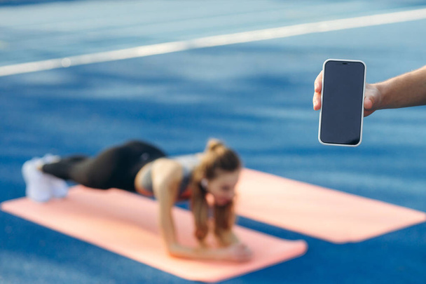 selective focus phone trainer holds phone in hand while girl stands in plank - Photo, Image