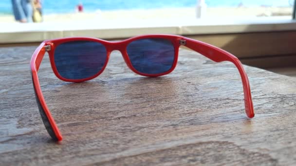Orange sunglasses on a table with a summer beach in the background - Πλάνα, βίντεο