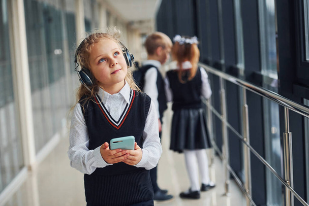 Little girl with phone and headphones standing if front of school kids in uniform that together in corridor. Conception of education. - Foto, Imagen