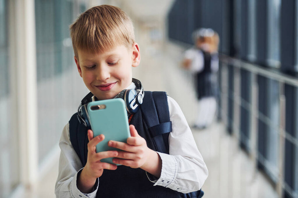 Little boywith phone and headphones standing if front of school kids in uniform that together in corridor. Conception of education. - Φωτογραφία, εικόνα
