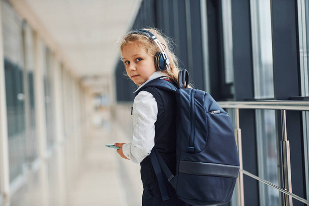 Cute little school girl in uniform with headphones and phone indoors. - Фото, изображение