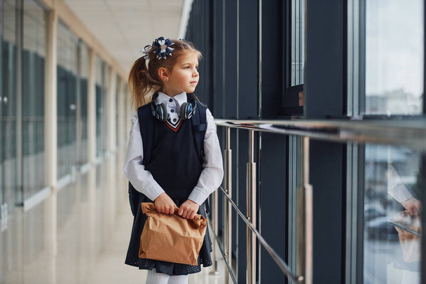 Young little school girl in uniform standing in hallway with package of dinner in hands. - Photo, Image