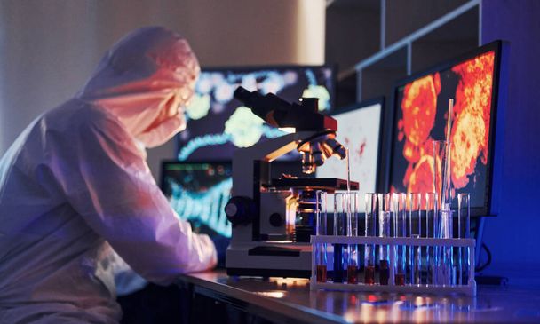 Monitors with information on the table. Scientist in white protective uniform works with coronavirus and blood tubes in laboratory. - Foto, Imagem