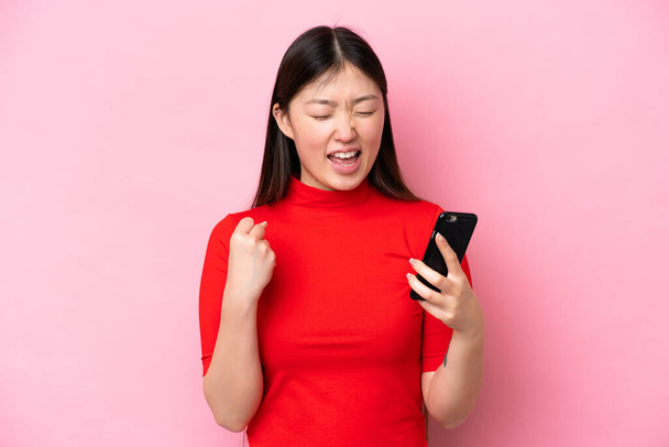 Young Chinese woman isolated on pink background with phone in victory position - Foto, afbeelding