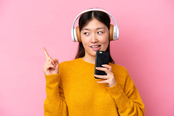 Young Chinese woman isolated on pink background listening music with a mobile and singing - Foto, Imagem