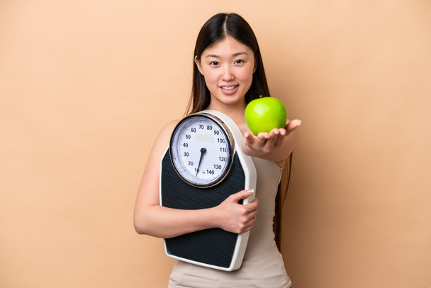Young Chinese woman isolated on beige background holding weighing machine and offering an apple - Фото, изображение
