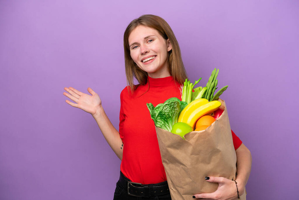 Young English woman holding a grocery shopping bag isolated on purple background extending hands to the side for inviting to come - Fotoğraf, Görsel