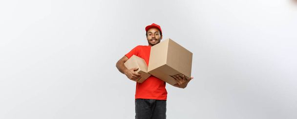 Αντίληψη παράδοσης - Πορτρέτο του Happy African American delivery man σε κόκκινο πανί κρατώντας ένα πακέτο κουτί. Απομονωμένος στο Grey studio Background. Αντιγραφή χώρου. - Φωτογραφία, εικόνα