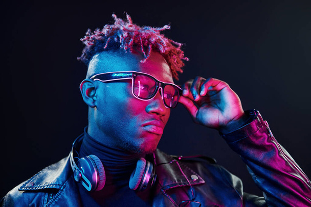 Stylish glasses and wireless headphones. Futuristic neon lighting. Young african american man in the studio. - Фото, изображение