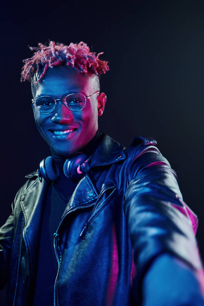 Posing with wireless headphones on neck. Futuristic neon lighting. Young african american man in the studio. - Photo, Image