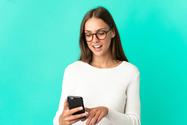 Молодая женщина на изолированном синем фоне отправляет сообщение или электронную почту с мобильного телефона - Фото, изображение