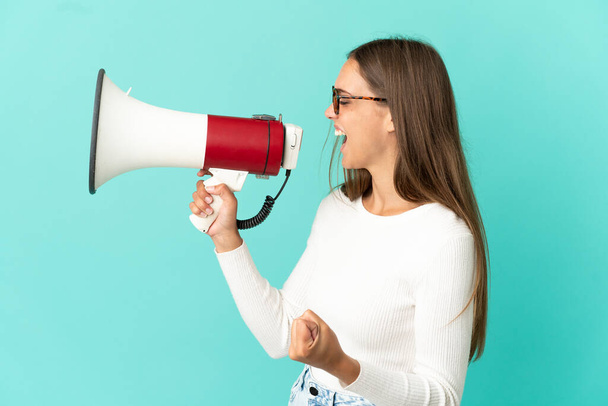 Young woman over isolated blue background shouting through a megaphone to announce something in lateral position - Photo, image