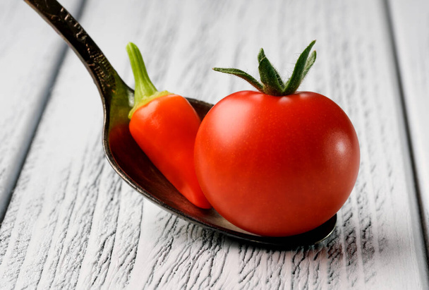 Small red ripe tomato with red hot pepper in a teaspoon on a light wooden background close-up - Photo, Image
