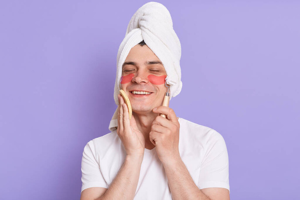 Indoor of joyful young man in towel use eye anti-wrinkle patches, holding sponge in hands and making face massage with roller, enjoying morning cosmetic procedures. - Foto, Imagen