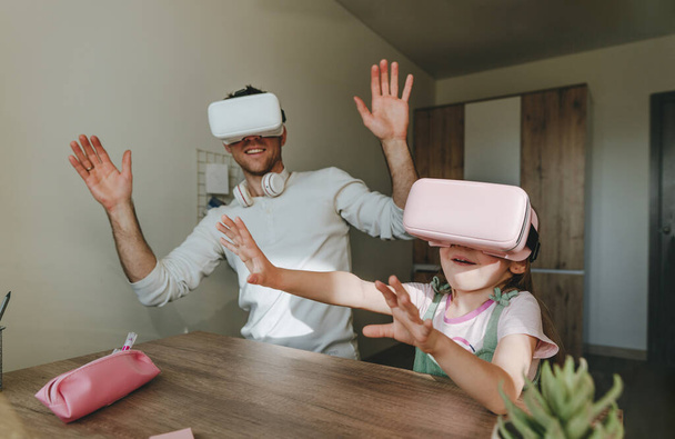 5 years old girl having fun time with her father using VR glasses at home for gaming or learning. Family activities concept. Modern technology using by family. Selective focus. - Фото, изображение