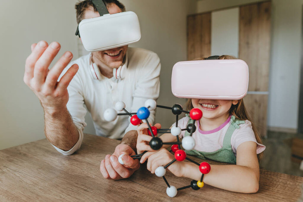 Little girl having fun time with her father using VR glasses at home for learning molecular structure. Homeschooling concept. Modern technology using by family. Selective focus. - Фото, изображение