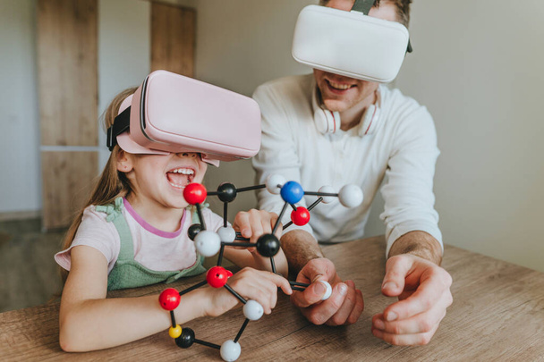 Little girl having fun time with her father using VR glasses at home for learning molecular structure. Homeschooling concept. Modern technology using by family. Selective focus. - Photo, image