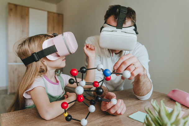 Το κοριτσάκι περνάει καλά με τον πατέρα της χρησιμοποιώντας γυαλιά εικονικής πραγματικότητας στο σπίτι για να μάθει μοριακή δομή. Φιλοξενία κατ 'οίκον. Σύγχρονη τεχνολογία που χρησιμοποιεί η οικογένεια. Επιλεκτική εστίαση. - Φωτογραφία, εικόνα