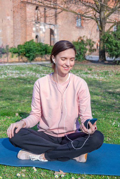Closeup portrait of beautiful woman sitting in lotus position on the grass in park and listening to music. - Foto, Bild