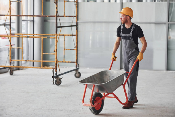 Walks with wheelbarrows. Man in grey uniform works indoors in modern big office at daytime. - Photo, Image