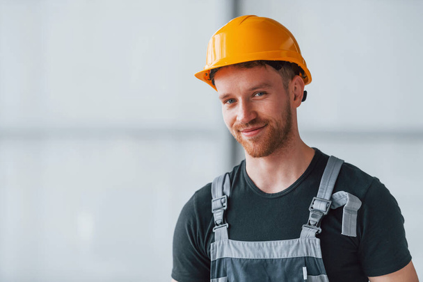 Portrait of man in grey uniform and orange hard hat that standing indoors in modern big office at daytime. - Foto, afbeelding