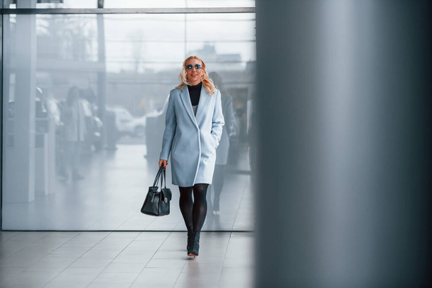 Beautiful blonde in elegant clothes with black handbag standing indoors against grey background. - Foto, Bild