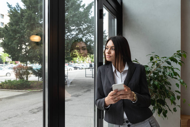 Young business woman in casual suit standing near window in the restaurant holding her smartphone waiting to waiter pack her lunch to go in the box. Freelancer expat waiting take away coffee or food - Fotó, kép