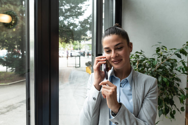 Young business woman in casual suit standing near window in the restaurant holding her smartphone waiting to waiter pack her lunch to go in the box. Freelancer expat waiting take away coffee or food - Foto, Imagem