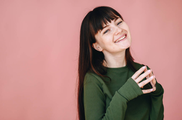 A pretty girl clasped her fingers and sincerely smiles on a pink background. Girl with bangs and freckles on her face - Foto, afbeelding