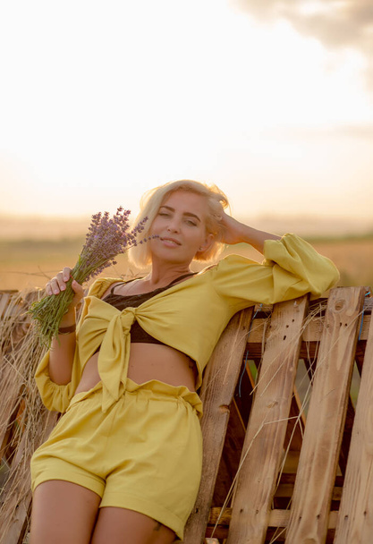 Pretty young woman in yellow among rural field with golden oat field on sunset background - Valokuva, kuva