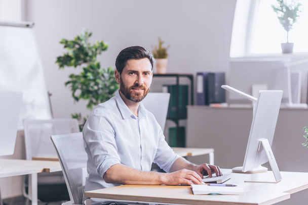Successful bearded freelance businessman looks at camera and smiles, portrait of broker in office works at computer - Foto, imagen