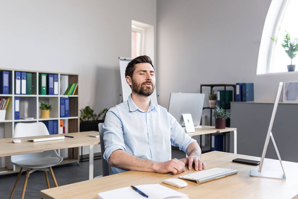 Businessman in the office with closed eyes performs breathing exercises, freelancer working on the computer - Foto, afbeelding