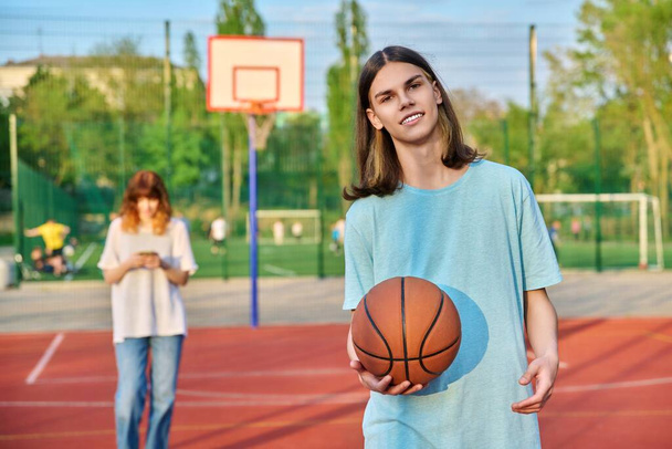 Portrait of young guy playing basketball on an outdoor game court, girl is out of focus. - Fotografie, Obrázek