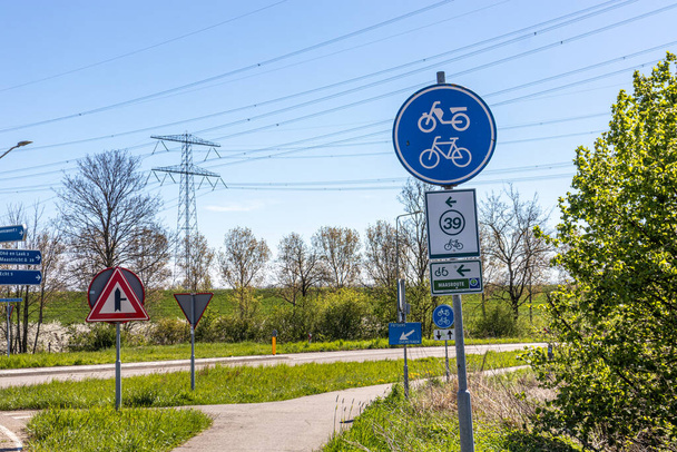 Different road sign: bike and moped lane, routes 39 and Maasrute,  caution triangle next crossroads, bike lane, country road, trees in background, sunny day in Stevensweert, South Limburg, Netherlands - Fotó, kép