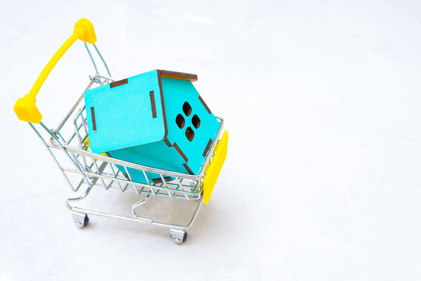 Miniature wooden house in a tiny shopping cart isolated on a neutral background. The concept of buying a house. - Фото, зображення