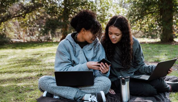 Close-up of two smiling teen sisters uses smartphone to surf the internet or chat with friends sitting at the park. - Foto, Imagem