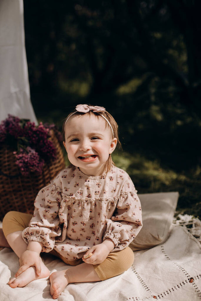 little baby girl sitting on a white picnic blanket in the garden. cheerful girl smiles broadly. child positively grimaces while sitting on a picnic. - Photo, Image