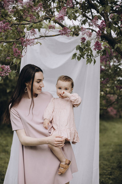 a young mother holds her daughter in her arms against a background of white cloth tied to a tree. young mother and baby girl near a white photo background in a lilac garden. happy mother and child. - Fotografie, Obrázek