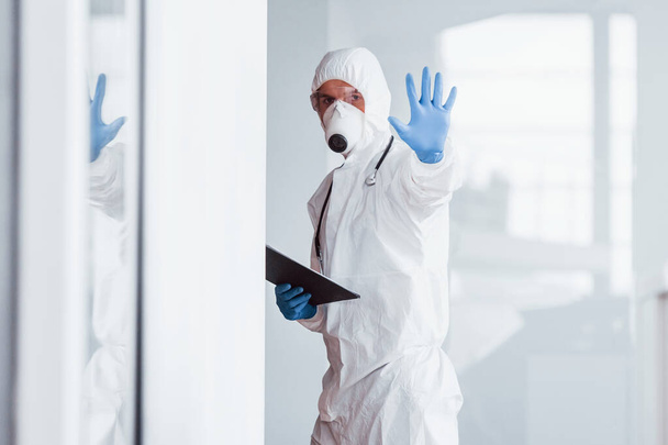 Shows stop gesture by hand. Male doctor scientist in lab coat, defensive eyewear and mask holding notepad in hands. - Photo, Image