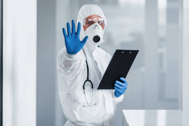 Shows stop gesture by hand. Male doctor scientist in lab coat, defensive eyewear and mask holding notepad in hands. - Фото, зображення