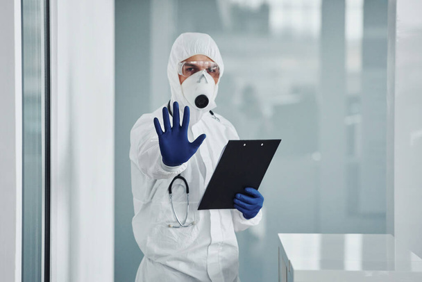 Shows stop gesture by hand. Male doctor scientist in lab coat, defensive eyewear and mask holding notepad in hands. - Valokuva, kuva