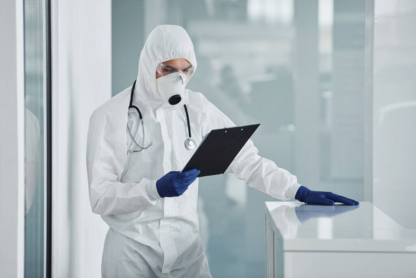 Male doctor scientist in lab coat, defensive eyewear and mask holding notepad in hands. - Photo, image