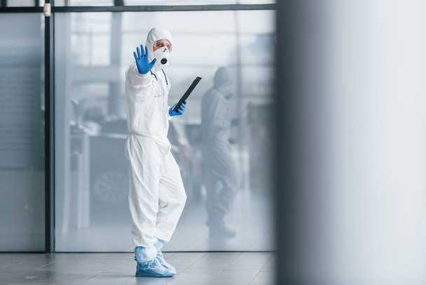 Shows stop gesture by hand. Male doctor scientist in lab coat, defensive eyewear and mask holding notepad in hands. - Fotó, kép